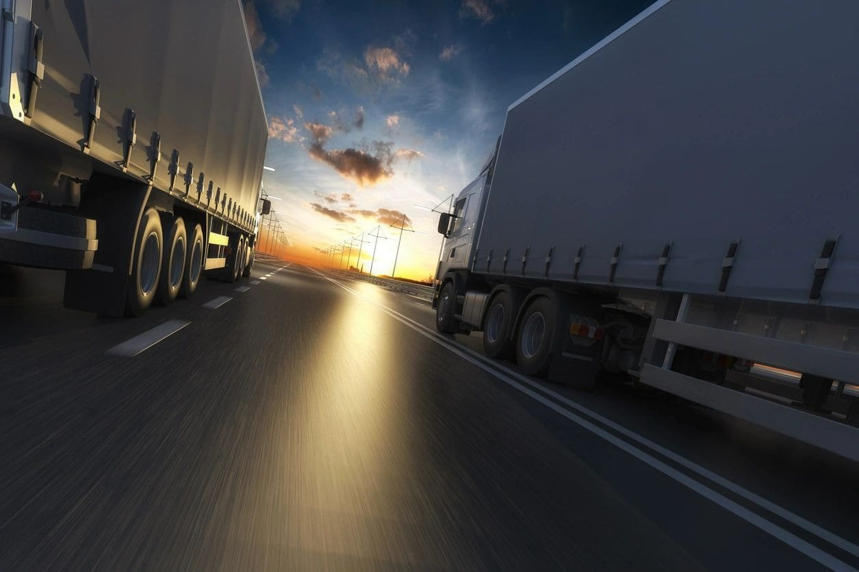 Two semi trucks driving down a highway at sunset.